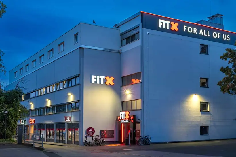Bild: Außenansicht eines FitX Fitnessstudios. Ein großes FitX-Logo mit orangefarbenem X ist zu sehen. Das Gebäude befindet sich in einem urbanen Umfeld.