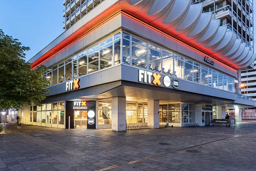 Bild: Außenansicht eines FitX Fitnessstudios. Ein großes FitX-Logo mit orangefarbenem X ist zu sehen. Das Gebäude befindet sich in einem urbanen Umfeld.