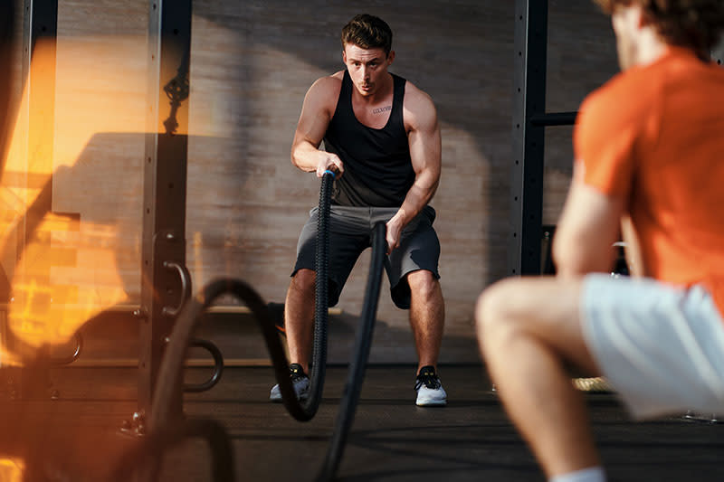 Bild: Ein Mann schlägt im functional-x-Kurs in der Turnecke Battle Ropes auf den Boden. Im Vordergrund macht ein anderer Teilnehmer Kniebeugen.