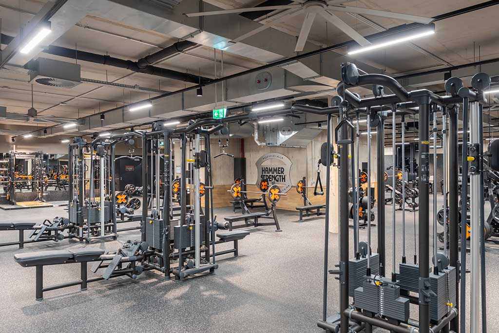 Weitläufiger Freihantelbereich mit verschiedenen Kraftgeräten im FitX Fitnessstudio Troisdorf-Mitte. An der Seite sind Kurzhanteln vor einer Spiegelwand aufgereiht. An der oberen Wand prangt der Schriftzug „FOR ALL OF US“.
