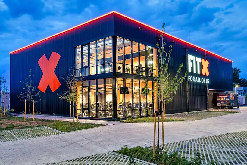 Bild: Außenansicht eines FitX Fitnessstudios. Ein großes FitX-Logo mit orangefarbenem X ist zu sehen. Das Gebäude befindet sich in einem urbanen Umfeld.