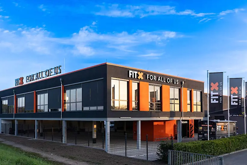 Bild: Außenansicht eines FitX Fitnessstudios. Ein großes FitX-Logo mit orangefarbenem X ist zu sehen. Das Gebäude befindet sich in einem urbanen Umfeld.