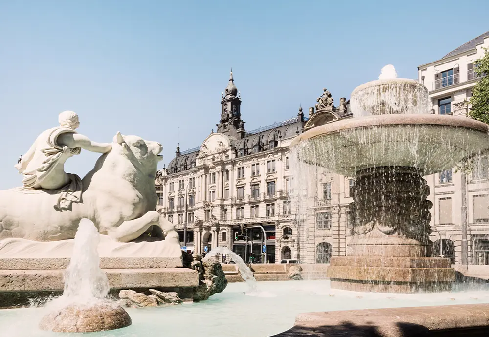 Bild: Blick auf den Wittelsbacher Brunnen zwischen Lenbachplatz und Maximiliansplatz, mit sprudelndem Wasser zwischen antiken Elementen und Gebäuden im Hintergrund.