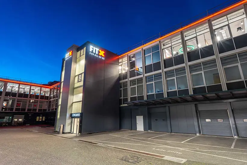 Bild: Außenansicht eines FitX Fitnessstudios. Ein großes FitX-Logo mit orangefarbenem X ist zu sehen. Das Gebäude befindet sich in einem urbanen Umfeld.