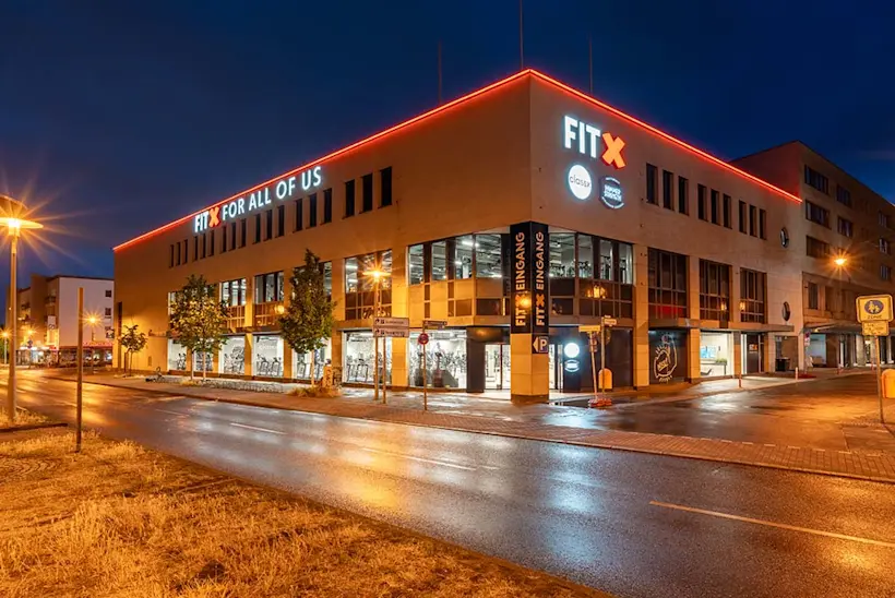Bild: Außenansicht eines FitX Fitnessstudios. Ein großes FitX-Logo mit orangefarbenem X ist zu sehen. Das Gebäude befindet sich in einem urbanen Umfeld.