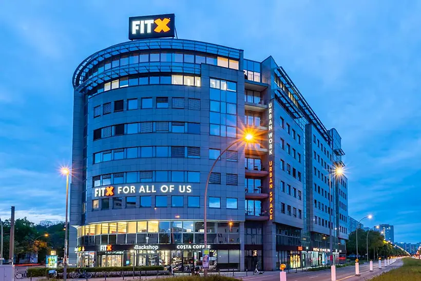Bild: Außenansicht eines FitX Fitnessstudios. Ein großes FitX-Logo mit orangefarbenem X ist zu sehen. Das Gebäude befindet sich in einem urbanen Umfeld.