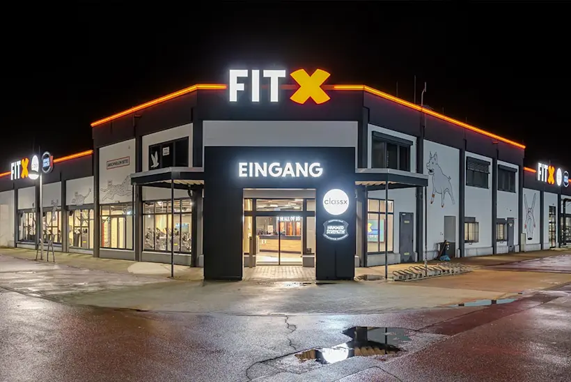 Bild: Außenansicht eines FitX Fitnessstudios. Ein großes FitX-Logo mit orangefarbenem X ist zu sehen. Das Gebäude befindet sich in einem urbanen Umfeld.