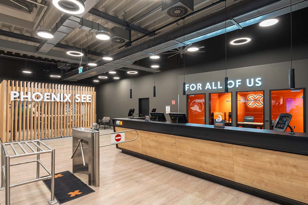 Empfangsbereich des FitX Fitnessstudios Dortmund-Phoenix See mit einer holzverkleideten Theke, einem Drehkreuz am Eingang und dem leuchtenden Schriftzug „FOR ALL OF US“ an der Wand.