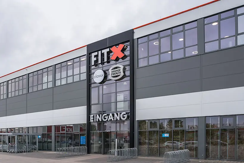 Bild: Außenansicht eines FitX Fitnessstudios. Ein großes FitX-Logo mit orangefarbenem X ist zu sehen. Das Gebäude befindet sich in einem urbanen Umfeld.