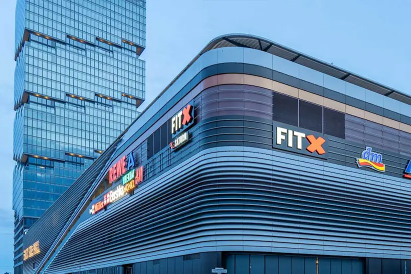 Bild: Außenansicht eines FitX Fitnessstudios. Ein großes FitX-Logo mit orangefarbenem X ist zu sehen. Das Gebäude befindet sich in einem urbanen Umfeld.