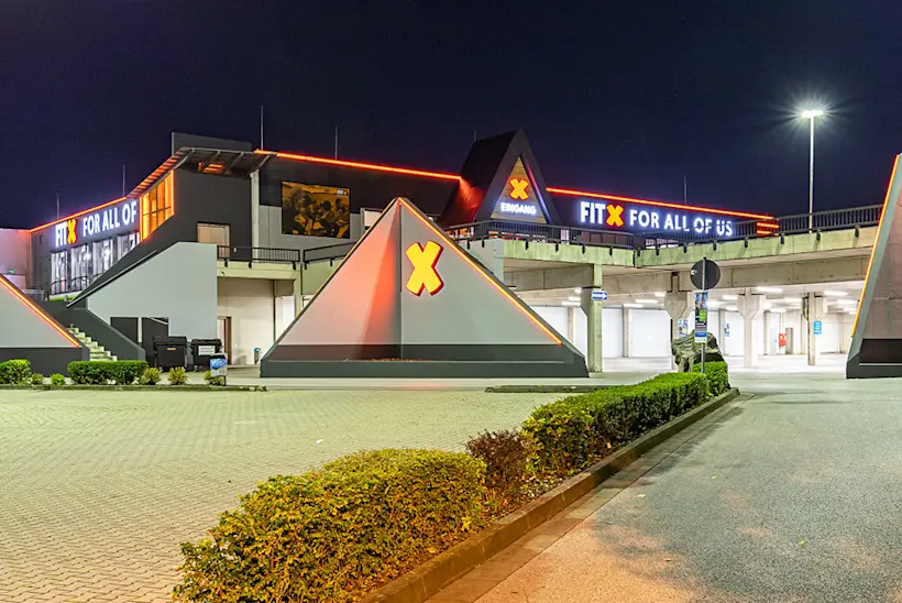 Bild: Außenansicht eines FitX Fitnessstudios. Ein großes FitX-Logo mit orangefarbenem X ist zu sehen. Das Gebäude befindet sich in einem urbanen Umfeld.