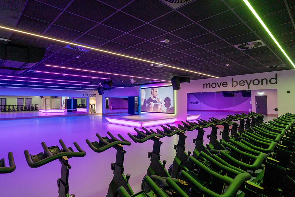 Großer, offener Kursraum im FitX Fitnessstudio Berlin-Moabit. An der hinteren Wand befindet sich eine erhöhte Bühne, dahinter eine Leinwand. Links und rechts davon sind Spiegelwände.