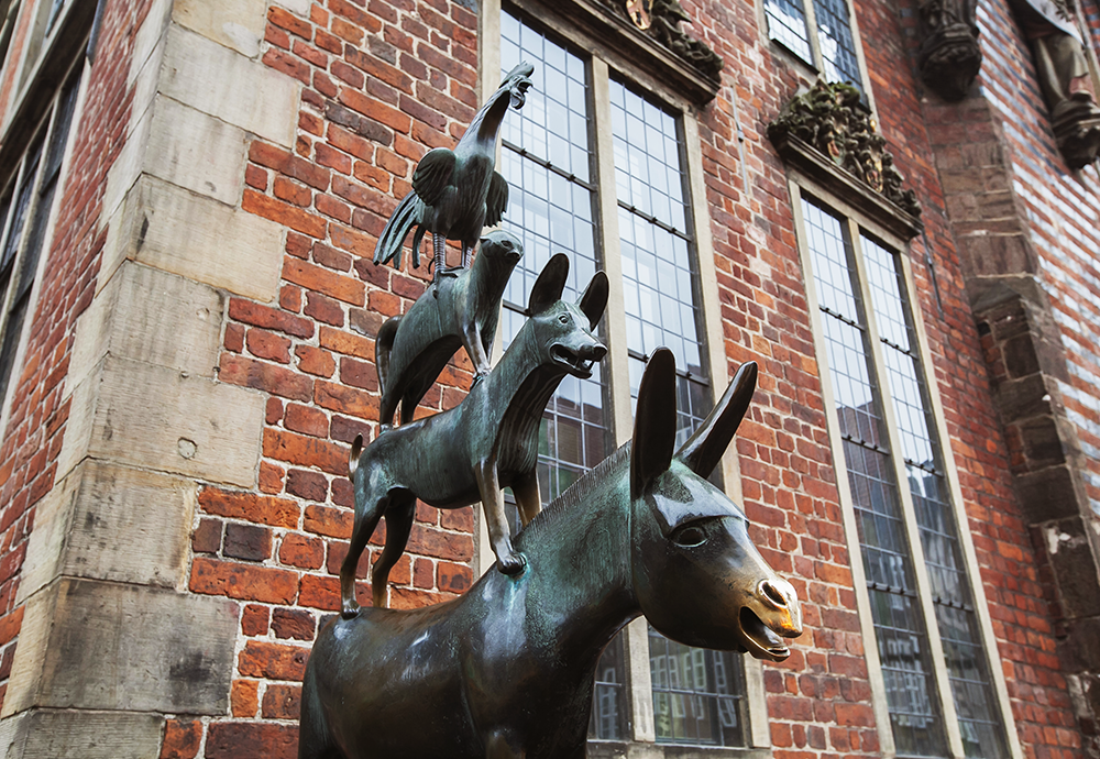 Bild: Die Statue der Bremer Stadtmusikanten aus Bronze steht vor einer Fassade aus roten Backsteinen mit hohen Sprossenfenstern.