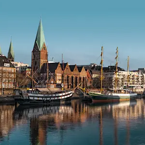 Zwei historische Segelschiffe liegen am Kai der Weser vor einer Kulisse aus roten Backsteinhäusern und Kirchen mit spitzen Türmen.