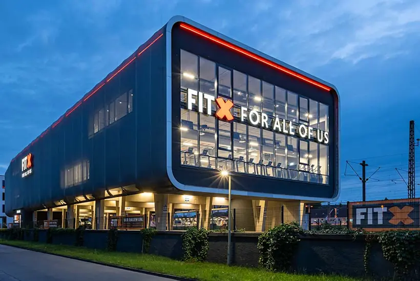 Bild: Außenansicht eines FitX Fitnessstudios. Ein großes FitX-Logo mit orangefarbenem X ist zu sehen. Das Gebäude befindet sich in einem urbanen Umfeld.