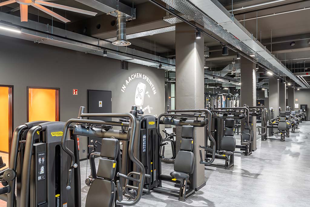 Gerätebereich im FitX Fitnessstudio Aachen-Mitte mit verschiedenen Kraftgeräten. Der Raum ist im industriellen Design gestaltet, mit offenen Deckenstrukturen und hellen Böden.