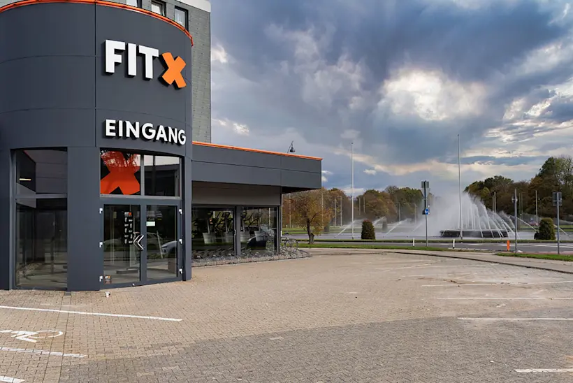 Bild: Außenansicht eines FitX Fitnessstudios. Ein großes FitX-Logo mit orangefarbenem X ist zu sehen. Das Gebäude befindet sich in einem urbanen Umfeld.