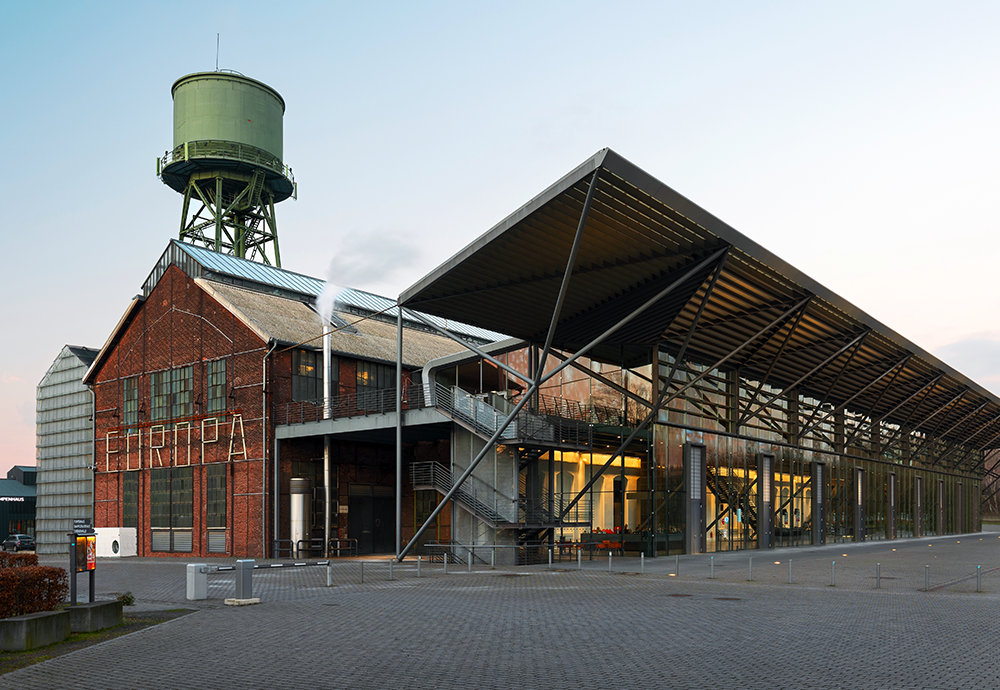 Bild: Altes Backsteingebäude mit dem Schriftzug „EUROPA“ und angrenzendem modernen Glasbau; im Hintergrund ein hoher, grüner Wasserturm – Teil der Jahrhunderthalle Bochum im Westpark.