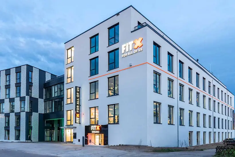 Bild: Außenansicht eines FitX Fitnessstudios. Ein großes FitX-Logo mit orangefarbenem X ist zu sehen. Das Gebäude befindet sich in einem urbanen Umfeld.