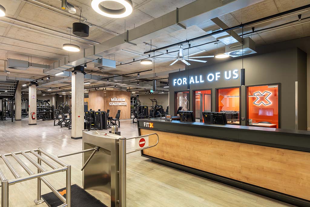 Empfangsbereich des FitX Fitnessstudios Mülheim-Stadtmitte mit einer holzverkleideten Theke, einem Drehkreuz am Eingang und dem leuchtenden Schriftzug „FOR ALL OF US“ an der Wand.