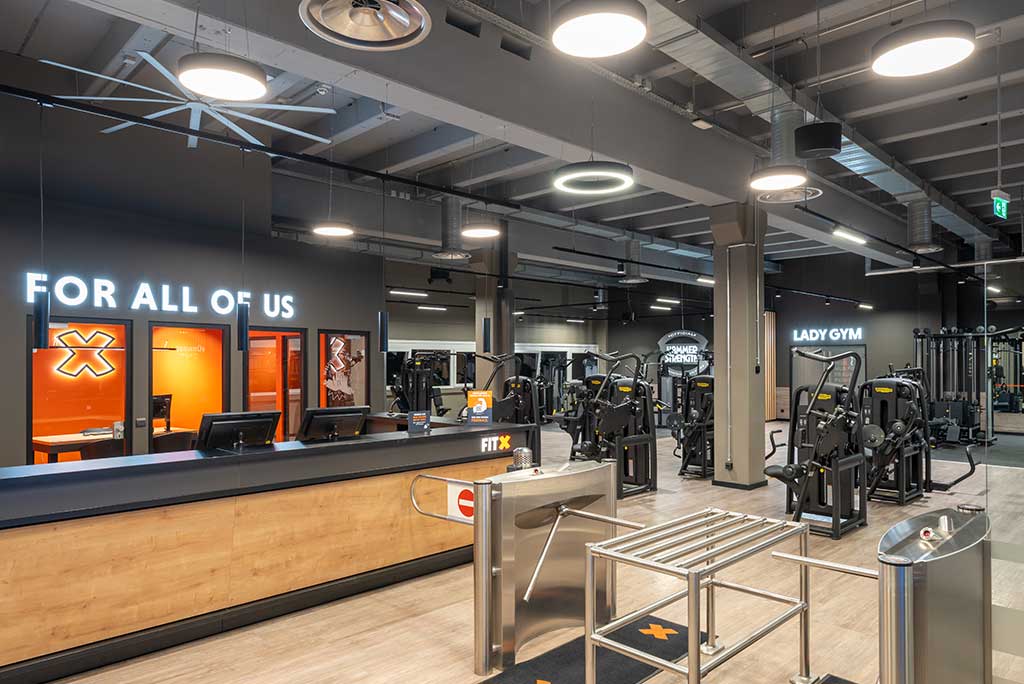 Empfangsbereich des FitX Fitnessstudios Nürnberg-Zabo mit einer holzverkleideten Theke, einem Drehkreuz am Eingang und dem leuchtenden Schriftzug „FOR ALL OF US“ an der Wand.
