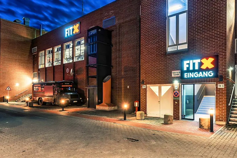 Bild: Außenansicht eines FitX Fitnessstudios. Ein großes FitX-Logo mit orangefarbenem X ist zu sehen. Das Gebäude befindet sich in einem urbanen Umfeld.