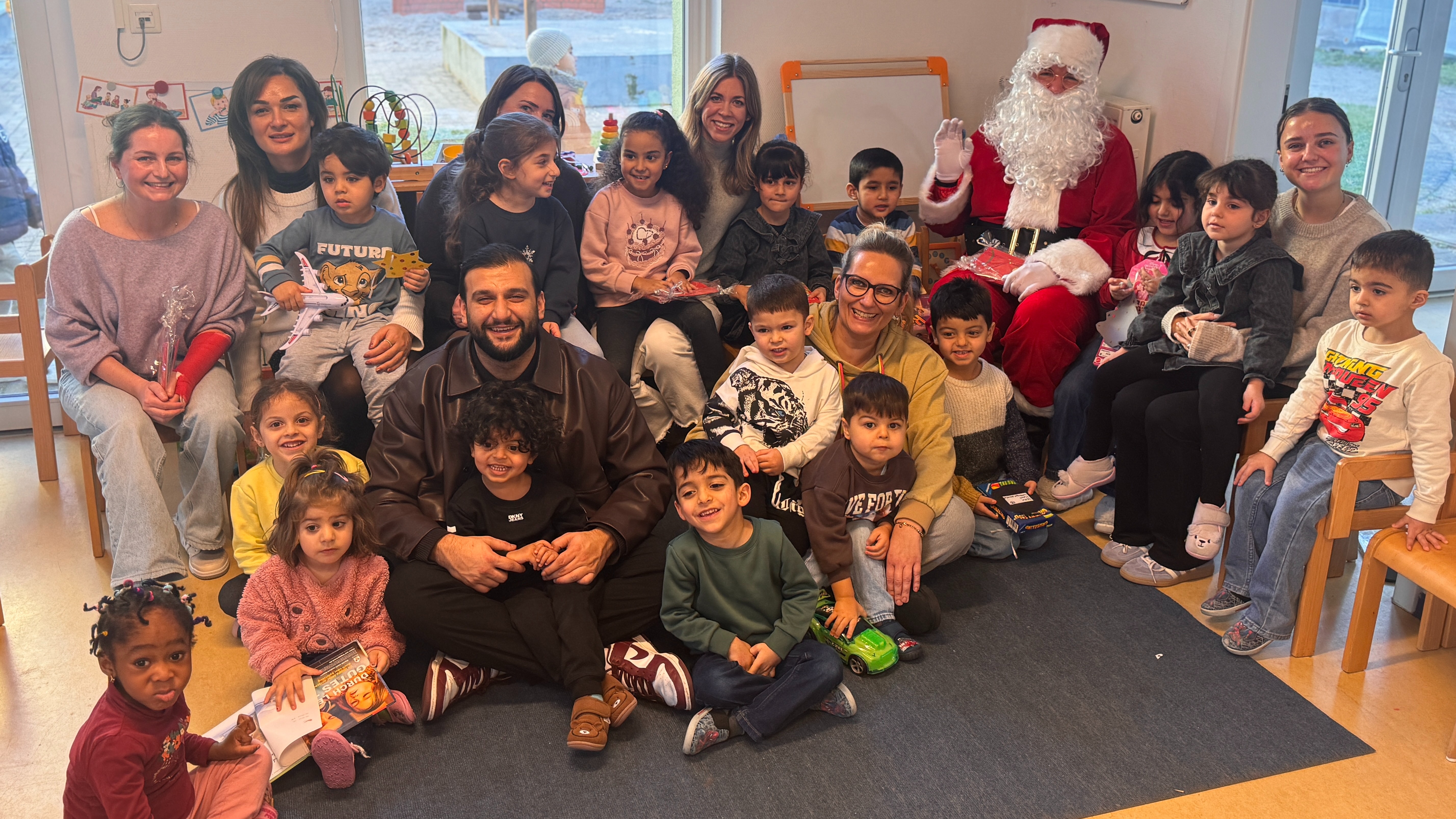 Bild: Einige FitX-Mitarbeitende haben die Geschenkübergabe der Wunschbaumaktion begleitet. Sie sitzen inmitten einer Kindergartengruppe, die vor Glück über ihre Geschenke strahlen. Ein Weihnachtsmann ist auch dabei.