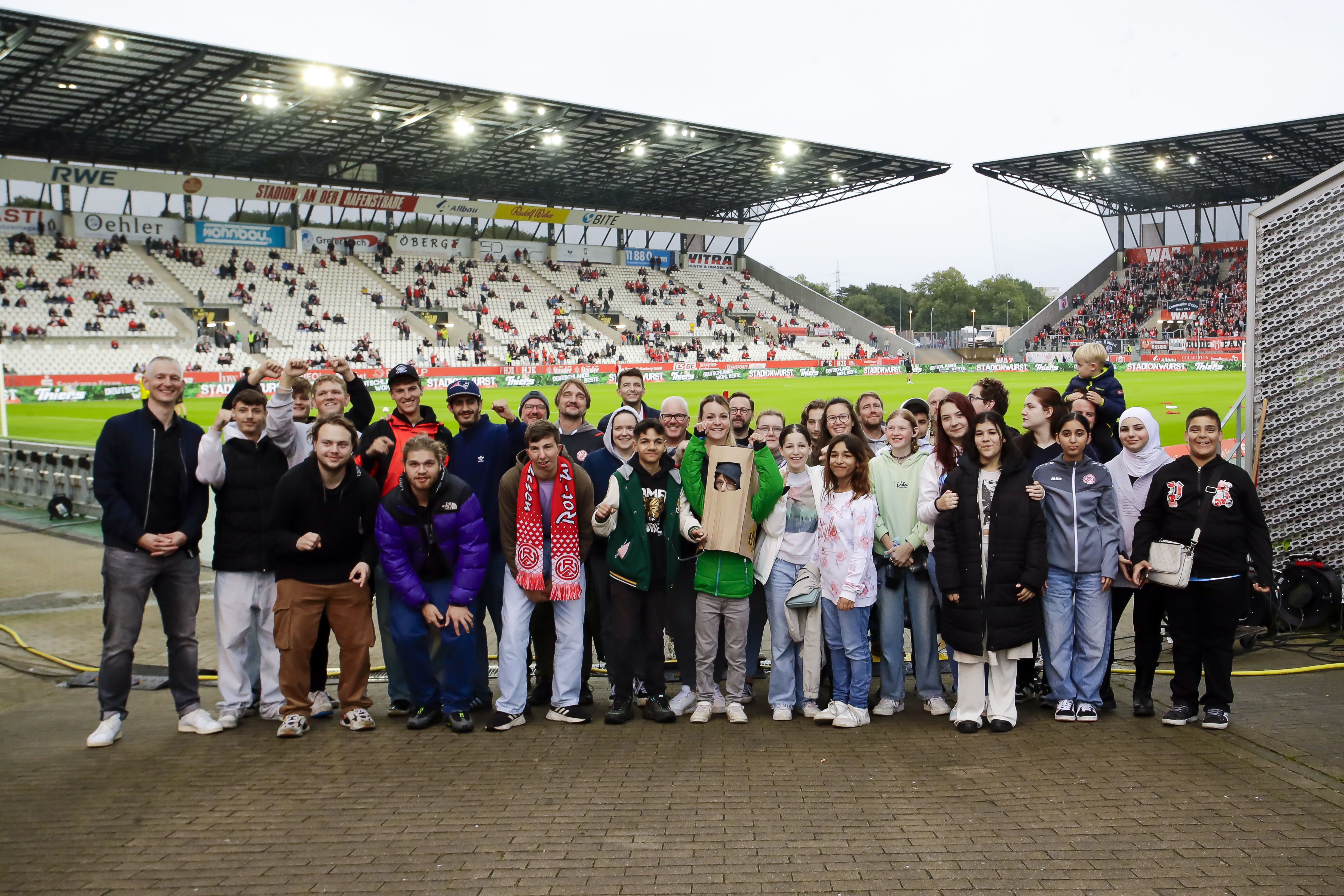 Bild: Eine Gruppe Menschen steht vor einem Fußballstadion. Im Hintergrund sieht man das Spielfeld und einige der Tribünen. Die Gruppe besteht weitestgehend aus Kindern und jungen Erwachsenen, links steht der FitX CEO Markus Vancraeyenest.