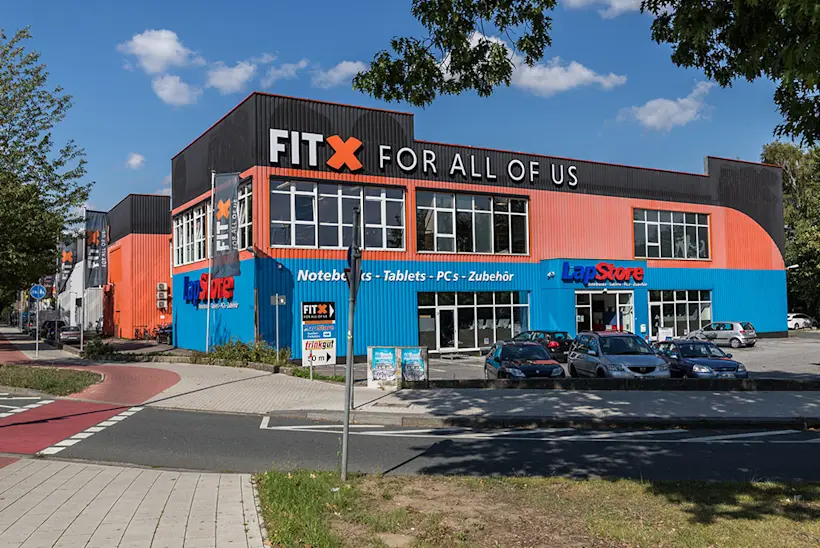 Bild: Außenansicht eines FitX Fitnessstudios. Ein großes FitX-Logo mit orangefarbenem X ist zu sehen. Das Gebäude befindet sich in einem urbanen Umfeld.