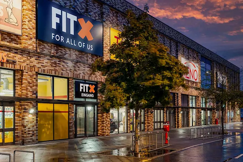 Bild: Außenansicht eines FitX Fitnessstudios. Ein großes FitX-Logo mit orangefarbenem X ist zu sehen. Das Gebäude befindet sich in einem urbanen Umfeld.