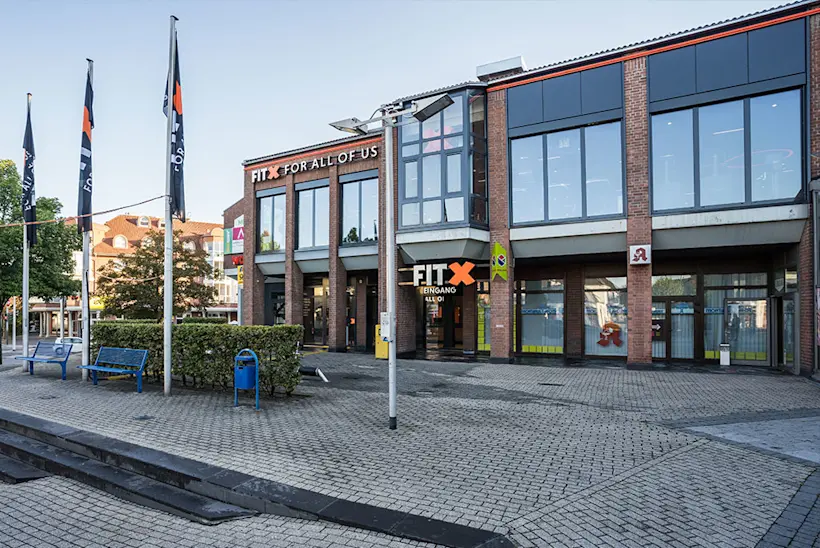 Bild: Außenansicht eines FitX Fitnessstudios. Ein großes FitX-Logo mit orangefarbenem X ist zu sehen. Das Gebäude befindet sich in einem urbanen Umfeld.