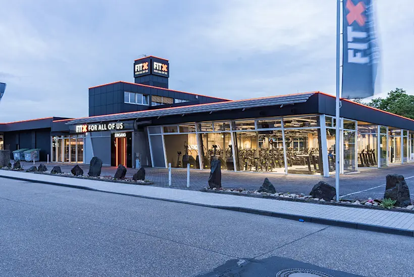 Bild: Außenansicht eines FitX Fitnessstudios. Ein großes FitX-Logo mit orangefarbenem X ist zu sehen. Das Gebäude befindet sich in einem urbanen Umfeld.