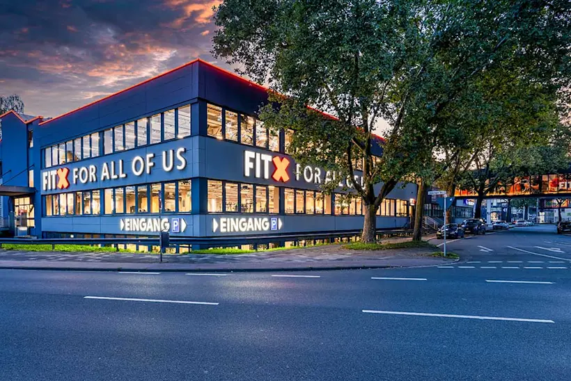 Bild: Außenansicht eines FitX Fitnessstudios. Ein großes FitX-Logo mit orangefarbenem X ist zu sehen. Das Gebäude befindet sich in einem urbanen Umfeld.