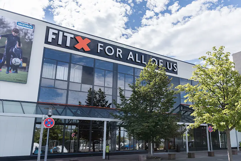 Bild: Außenansicht eines FitX Fitnessstudios. Ein großes FitX-Logo mit orangefarbenem X ist zu sehen. Das Gebäude befindet sich in einem urbanen Umfeld.