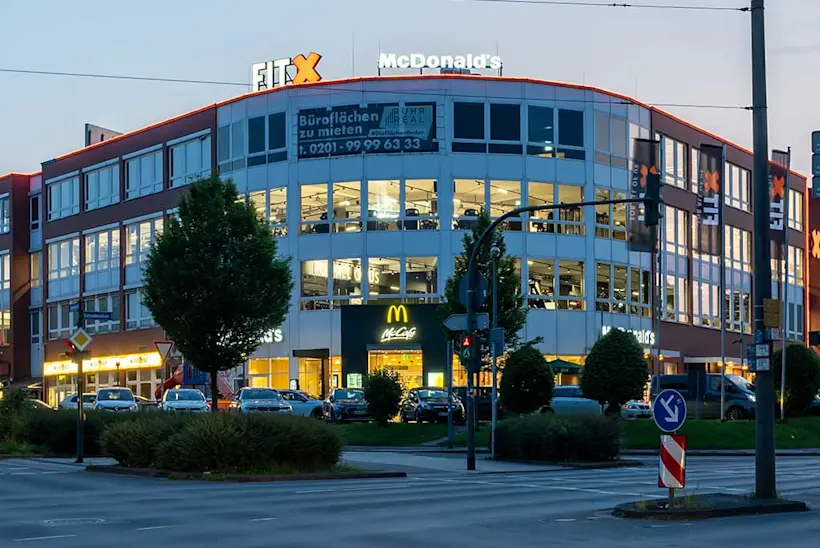 Bild: Außenansicht eines FitX Fitnessstudios. Ein großes FitX-Logo mit orangefarbenem X ist zu sehen. Das Gebäude befindet sich in einem urbanen Umfeld.
