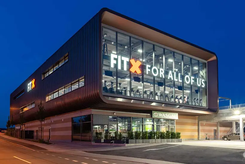 Bild: Außenansicht eines FitX Fitnessstudios. Ein großes FitX-Logo mit orangefarbenem X ist zu sehen. Das Gebäude befindet sich in einem urbanen Umfeld.
