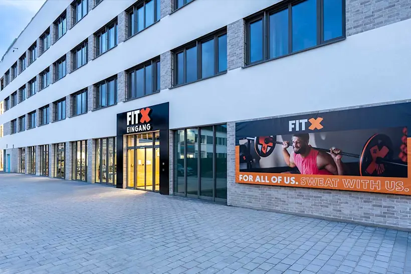 Bild: Außenansicht eines FitX Fitnessstudios. Ein großes FitX-Logo mit orangefarbenem X ist zu sehen. Das Gebäude befindet sich in einem urbanen Umfeld.