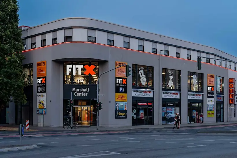 Bild: Außenansicht eines FitX Fitnessstudios. Ein großes FitX-Logo mit orangefarbenem X ist zu sehen. Das Gebäude befindet sich in einem urbanen Umfeld.