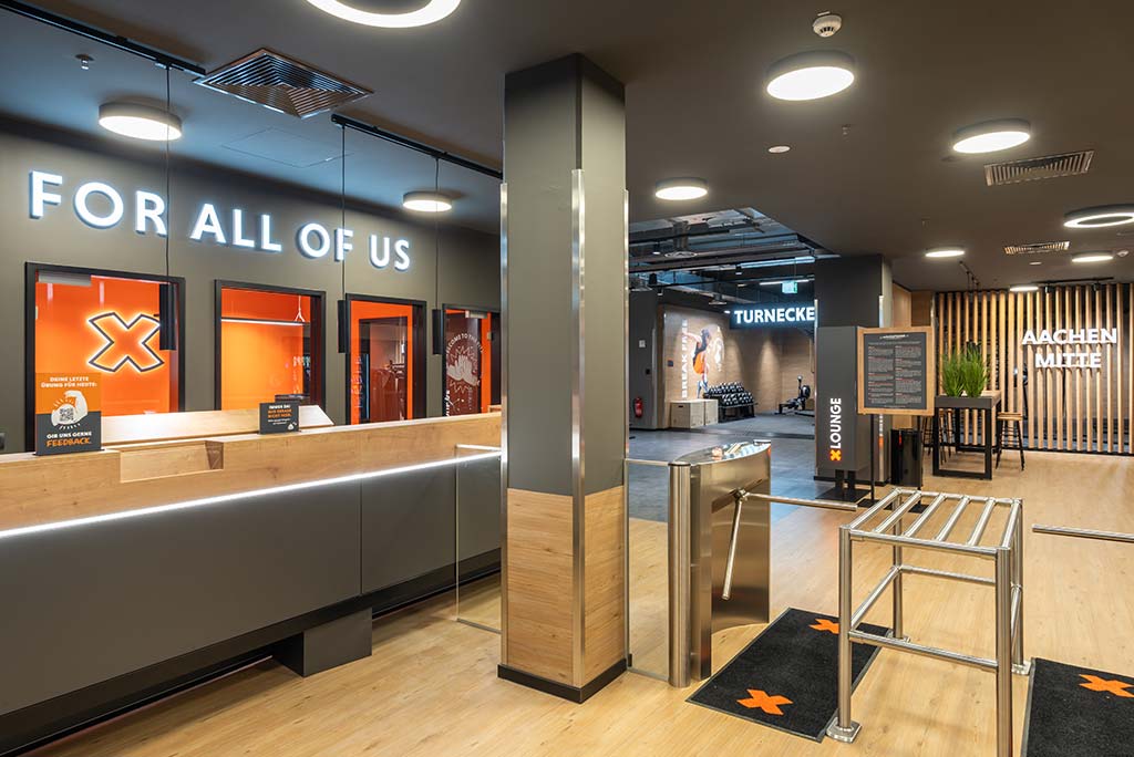 Empfangsbereich des FitX Fitnessstudios Aachen-Mitte mit einer holzverkleideten Theke, einem Drehkreuz am Eingang und dem leuchtenden Schriftzug „FOR ALL OF US“ an der Wand.