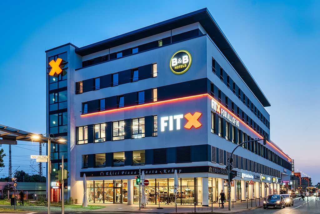 Außenansicht des FitX Fitnessstudios Troisdorf-Mitte. Ein großes FitX-Logo mit orangefarbenem X ist zu sehen. Das Gebäude befindet sich in einem urbanen Umfeld.
