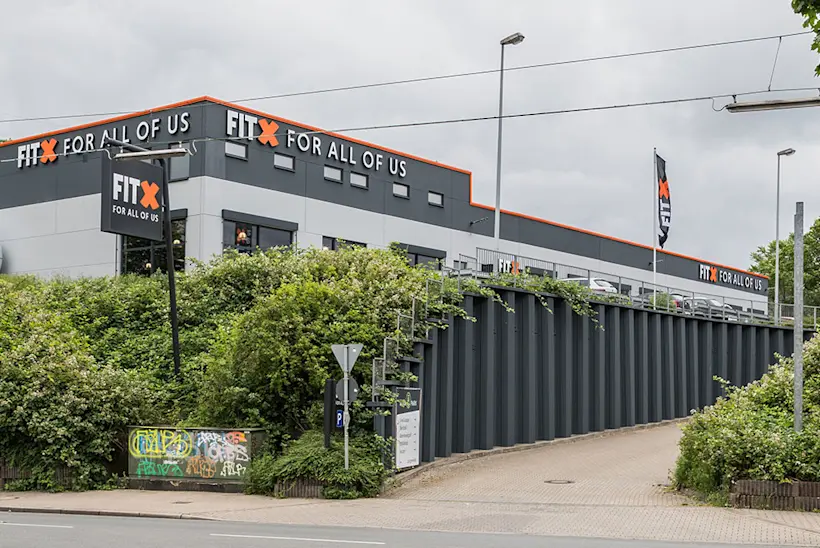 Bild: Außenansicht eines FitX Fitnessstudios. Ein großes FitX-Logo mit orangefarbenem X ist zu sehen. Das Gebäude befindet sich in einem urbanen Umfeld.
