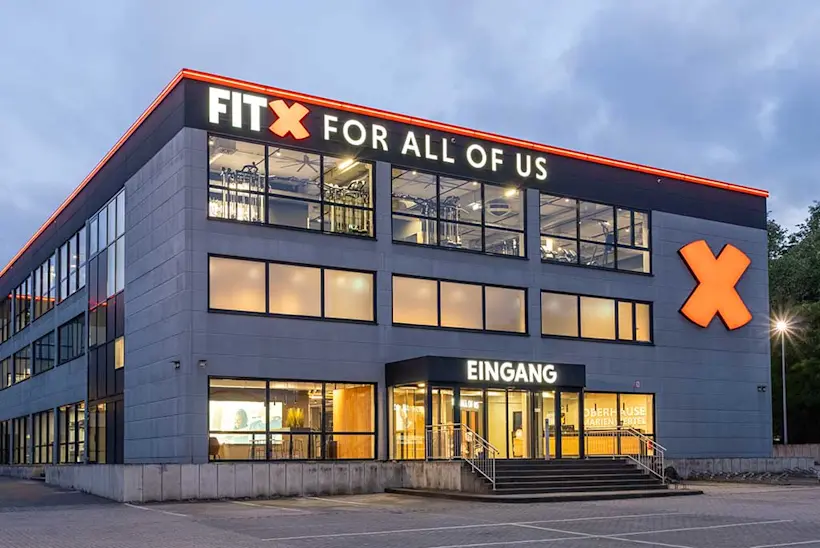Bild: Außenansicht eines FitX Fitnessstudios. Ein großes FitX-Logo mit orangefarbenem X ist zu sehen. Das Gebäude befindet sich in einem urbanen Umfeld.