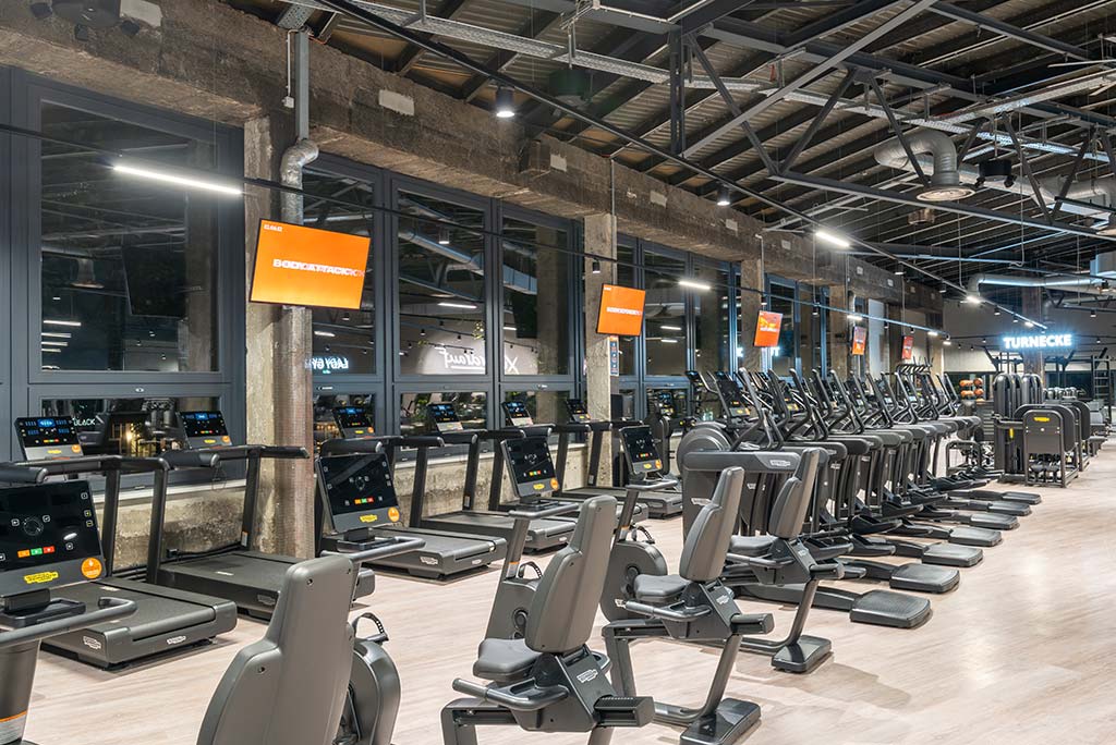 Offener Bereich für Ausdauertraining im FitX Fitnessstudio Berlin-Ku'damm mit verschiedenen Cardio-Geräten. An den Wänden hängen Fernseher.