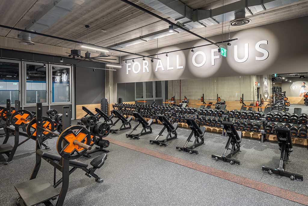 Weitläufiger Freihantelbereich mit verschiedenen Kraftgeräten im FitX Fitnessstudio Leipzig-Neulindenau. An der Seite sind Kurzhanteln vor einer Spiegelwand aufgereiht. An der oberen Wand prangt der Schriftzug „FOR ALL OF US“.