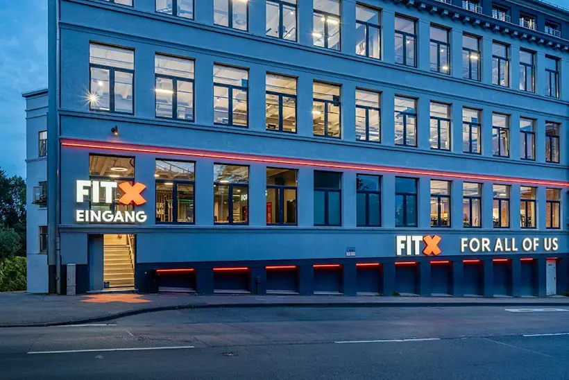 Bild: Außenansicht eines FitX Fitnessstudios. Ein großes FitX-Logo mit orangefarbenem X ist zu sehen. Das Gebäude befindet sich in einem urbanen Umfeld.