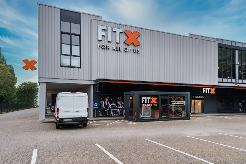 Bild: Außenansicht eines FitX Fitnessstudios. Ein großes FitX-Logo mit orangefarbenem X ist zu sehen. Das Gebäude befindet sich in einem urbanen Umfeld.