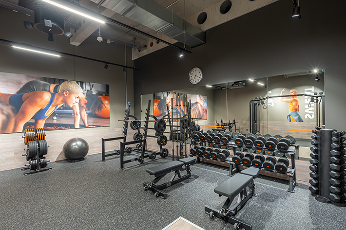 Empfangsbereich des FitX Fitnessstudios Augsburg-Lechhausen mit einer holzverkleideten Theke, einem Drehkreuz am Eingang und dem leuchtenden Schriftzug „FOR ALL OF US“ an der Wand.