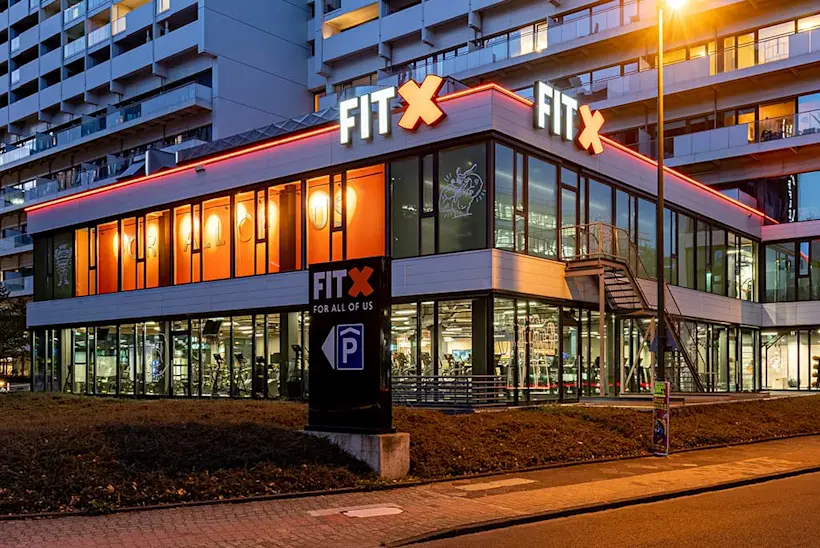 Bild: Außenansicht eines FitX Fitnessstudios. Ein großes FitX-Logo mit orangefarbenem X ist zu sehen. Das Gebäude befindet sich in einem urbanen Umfeld.