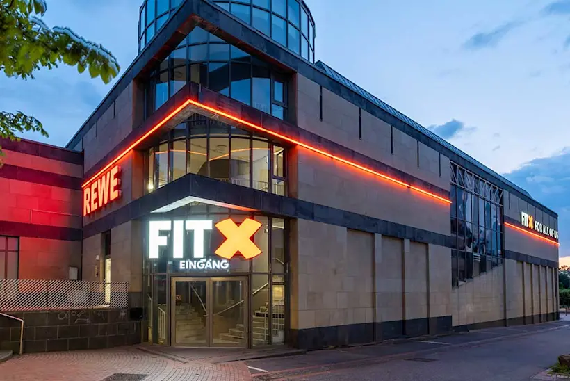 Bild: Außenansicht eines FitX Fitnessstudios. Ein großes FitX-Logo mit orangefarbenem X ist zu sehen. Das Gebäude befindet sich in einem urbanen Umfeld.