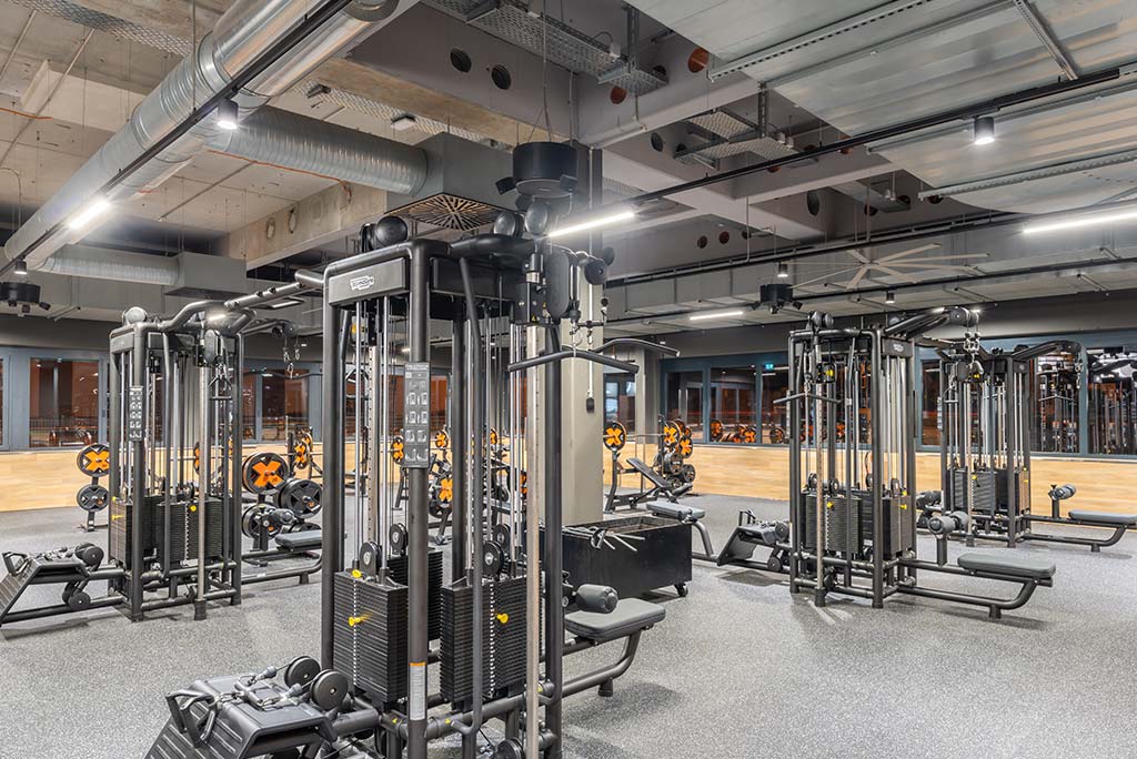 Weitläufiger Freihantelbereich mit verschiedenen Kraftgeräten im FitX Fitnessstudio Augsburg-Lechhausen. An der Seite sind Kurzhanteln vor einer Spiegelwand aufgereiht. An der oberen Wand prangt der Schriftzug „FOR ALL OF US“.